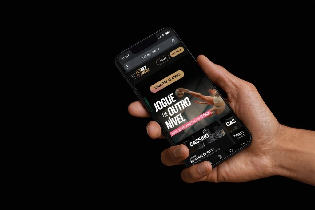 Uma mão segurando um smartphone que exibe a página inicial do site BetMGM. Na tela, destaca-se o botão dourado 'Registrar' no topo e um banner central com o texto 'Cadastre-se agora', incentivando o BetMGM cadastro para apostas esportivas e cassino.