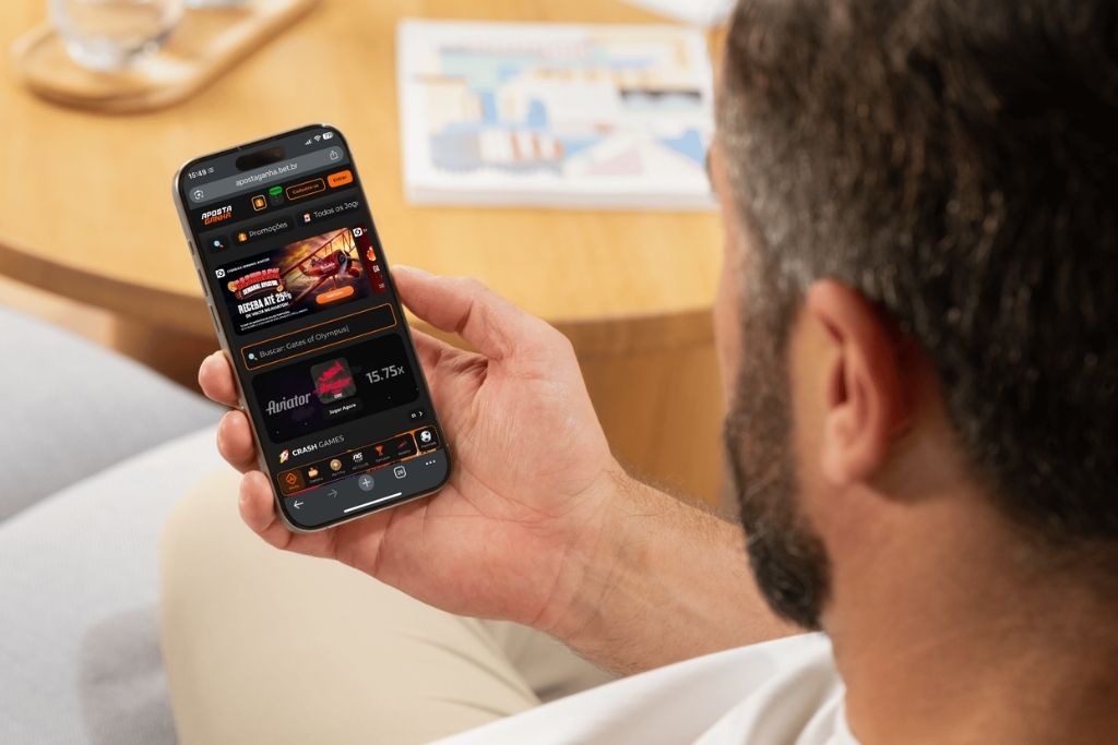 Homem segurando um smartphone que exibe a página inicial do site para realizar o cadastro no Aposta Ganha enquanto relaxa em uma sala.