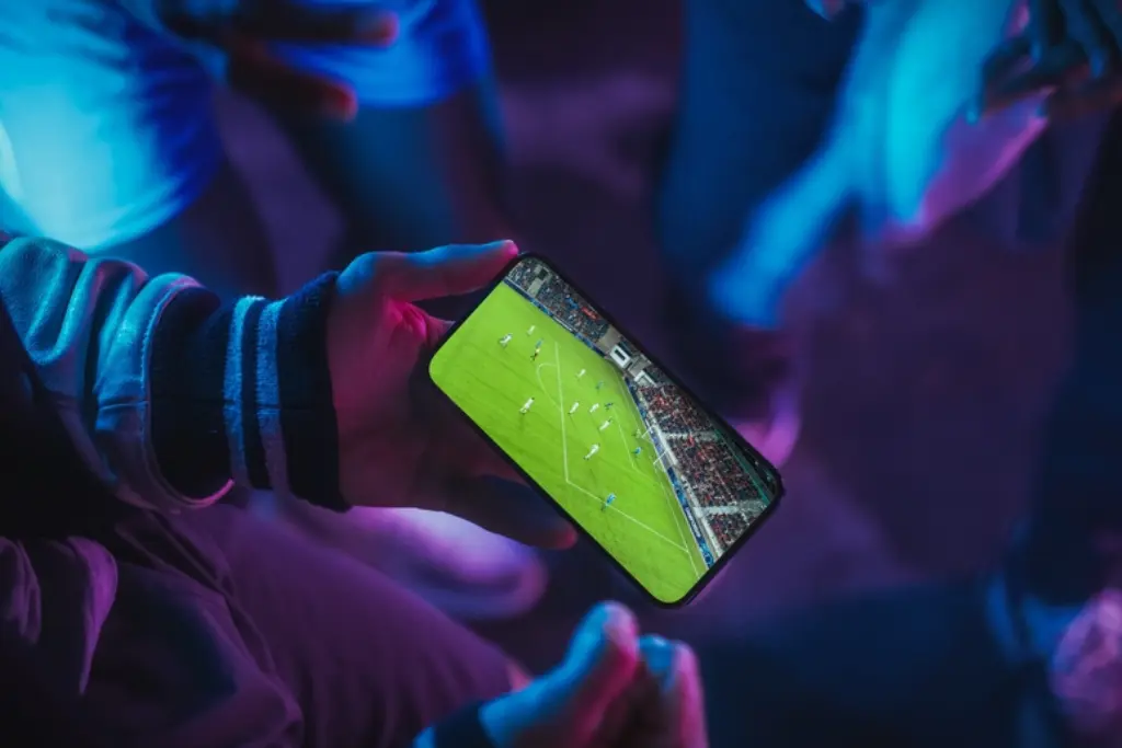 Close-up das mãos de uma pessoa segurando um smartphone que transmite uma partida de futebol ao vivo, representando o acesso a casas de apostas autorizadas em um ambiente com iluminação neon azul e roxa.