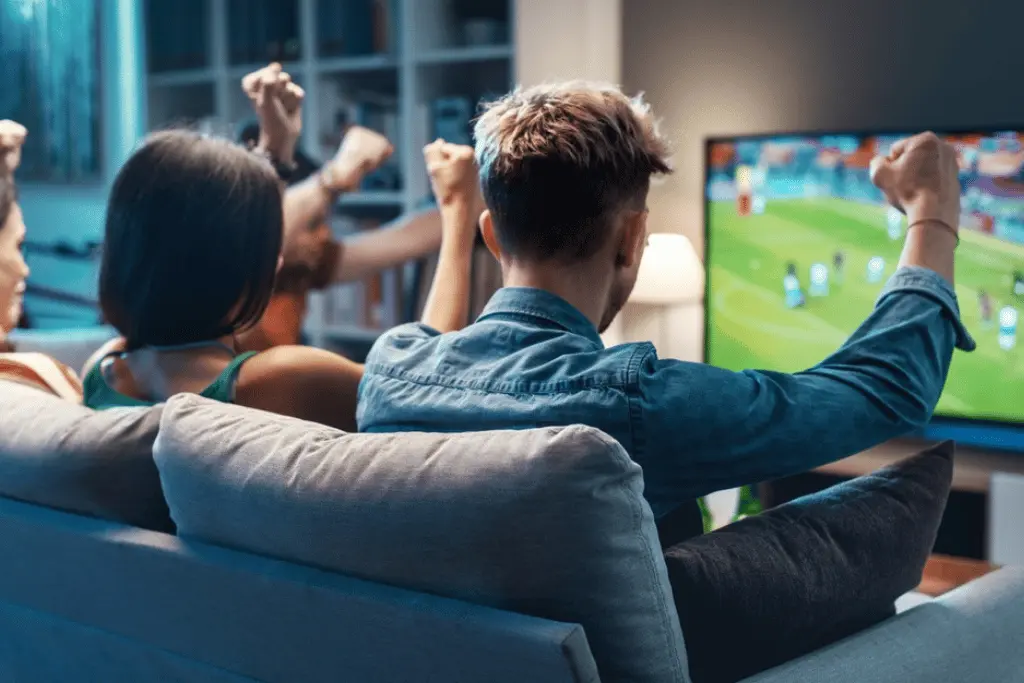 Grupo de amigos de costas, sentados em um sofá, comemorando com os braços levantados enquanto assistem a um jogo de futebol na TV, ilustrando o entretenimento seguro oferecido pelas casas de apostas autorizadas.