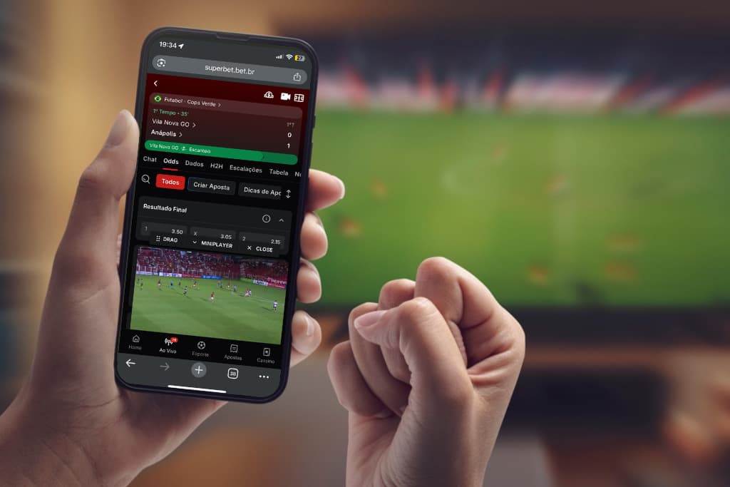 Close-up das mãos de uma pessoa segurando um smartphone com um aplicativo de apostas esportivas aberto. A tela mostra uma partida de futebol entre Vila Nova e Anápolis com odds em tempo real. Ao fundo, uma TV desfocada exibe o jogo, ilustrando como apostar ao vivo pelo celular durante a transmissão.