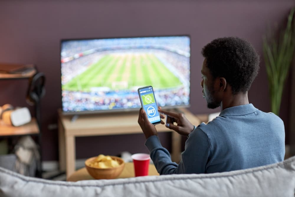 Um homem sentado em um sofá assistindo a uma partida de futebol em uma TV de tela plana enquanto utiliza um aplicativo de apostas no smartphone. Sobre a mesa, há petiscos e uma bebida. A cena demonstra o ambiente doméstico de como apostar ao vivo acompanhando os lances do jogo.