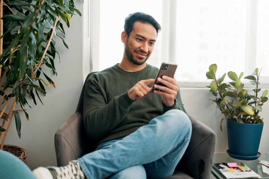 Um homem jovem sorridente, vestindo uma camisola verde e calças de ganga, sentado confortavelmente numa poltrona enquanto utiliza o telemóvel. Ele parece navegar em sites de entretenimento ou procurar por casas de apostas com bónus grátis num ambiente doméstico iluminado e decorado com plantas.