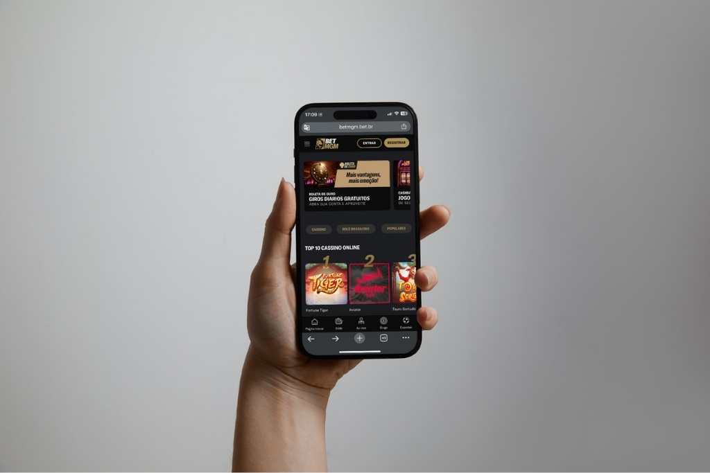 Uma mão segurando um smartphone que exibe a interface de uma plataforma de jogos online. O dispositivo mostra opções de cassino e promoções típicas de casas de apostas com bónus grátis, com um design moderno em tons de preto e dourado sobre um fundo cinza neutro.