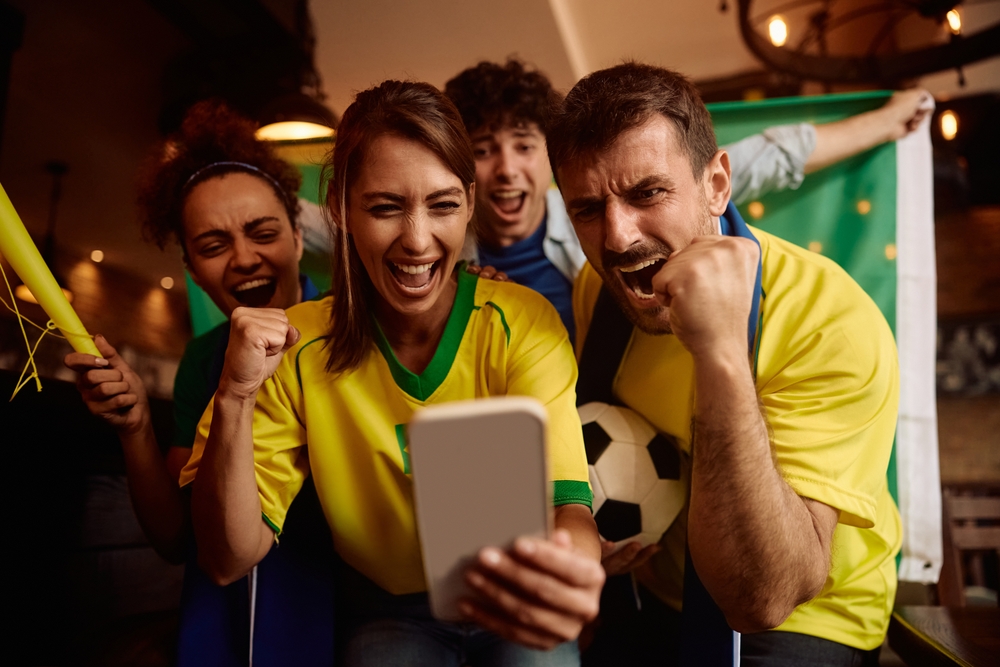Grupo de torcedores brasileiros comemorando enquanto acompanha uma partida pelo celular, com bandeira do Brasil e clima de emoção, representando a experiência de apostar nos favoritos da Copa do Mundo.