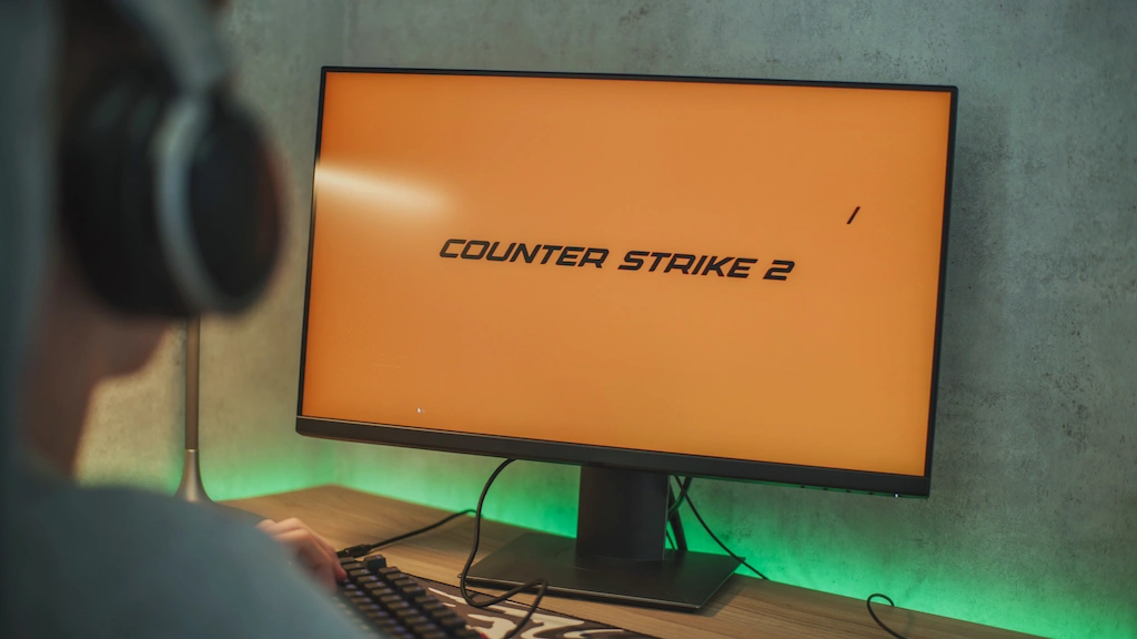 Monitor exibindo o logotipo 'Counter Strike 2' em tela laranja, focado em apostas CS2, com jogador de fones de ouvido e teclado em primeiro plano.