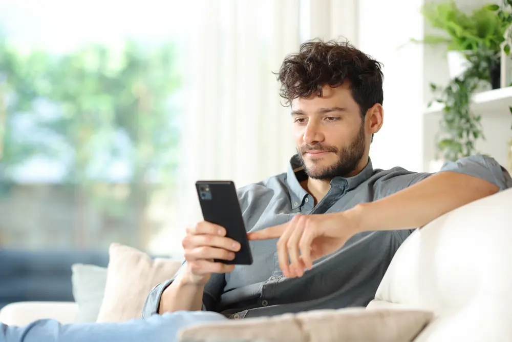 Homem usando o celular no sofá enquanto pesquisa o melhor site de apostas para iniciantes.